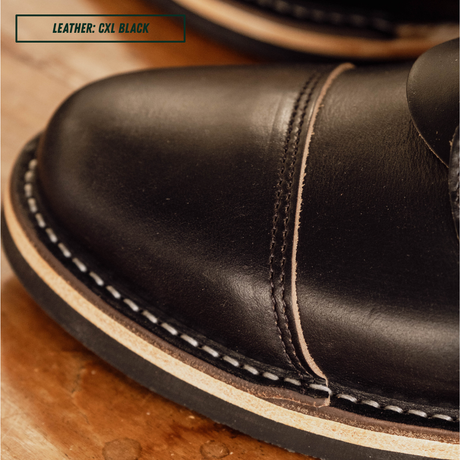 Close-up of a black leather shoe toe with contrast stitching and a light tan welt, photographed on a wooden surface. A text label in the top left reads 'LEATHER: CXL BLACK'.