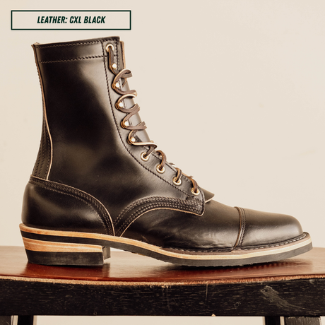 A single black leather lace-up ankle boot shown in side profile, resting on a wooden surface against a plain light beige background. A small label at the top reads 'LEATHER: CXL BLACK'.