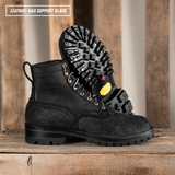 A pair of black leather lace-up boots with rugged lug soles, displayed on a rustic wooden surface with one boot standing upright and the other lying on its side to show the sole. A text overlay in the top left reads 'LEATHER: MAX SUPPORT BLACK'.