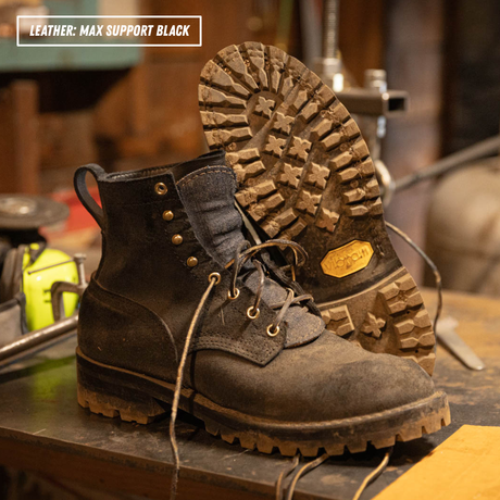 A pair of rugged lace-up work boots in dark brown leather, with one boot standing upright and the other lying on its side to show the sole. The boots are placed on a workbench in a workshop setting.