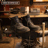 A pair of black leather lace-up work boots displayed on an anvil in a rustic workshop setting. A text overlay in the top-left corner reads 'LEATHER: MAX SUPPORT BLACK'.