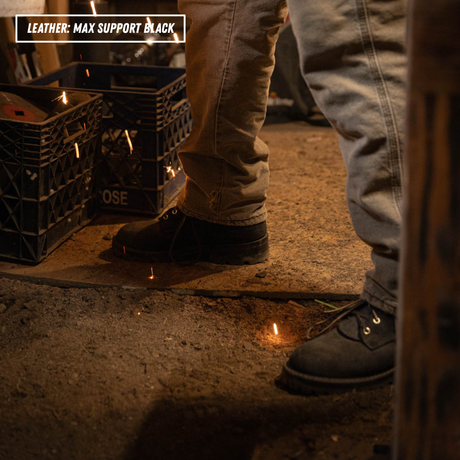 A close-up lifestyle shot of a person wearing black leather work boots with tan pants, standing on a concrete floor in a workshop. Sparks are visible on the floor around the boots, and a text overlay in the top-left corner reads 'LEATHER: MAX SUPPORT BLACK'.