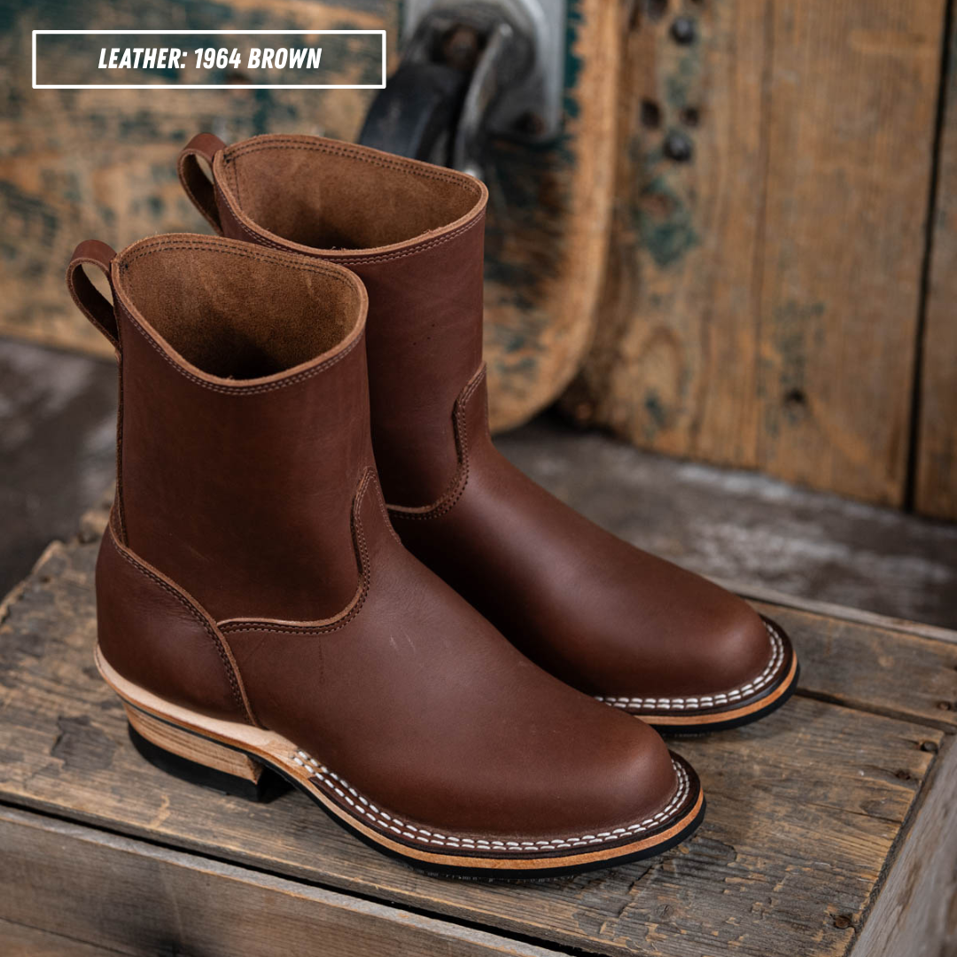 A pair of brown leather mid-calf boots with white contrast stitching and stacked heels, displayed on a rustic wooden crate with a wooden background. A text overlay in the top left reads 'LEATHER: 1964 BROWN'.