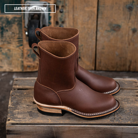 A pair of brown leather mid-calf boots with pull tabs and stacked low heels, displayed on rustic wooden planks with a weathered wood background. A text overlay in the top left reads 'LEATHER: 1964 BROWN'.