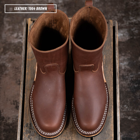 A pair of brown leather mid-calf boots with white contrast stitching, displayed on a rustic wooden crate. A text overlay in the top left corner reads 'LEATHER: 1964 BROWN'.