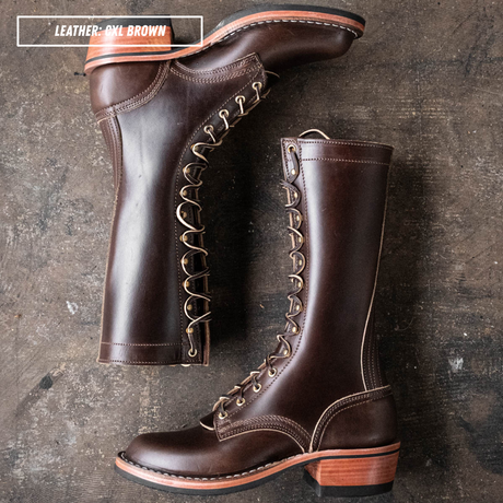 Two dark brown leather lace-up boots displayed on a textured dark surface, with one boot standing upright and the other lying on its side to show the side profile and heel.