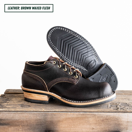 A pair of dark brown waxed leather lace-up boots, with one boot standing upright and the other tilted to show the treaded outsole. They are displayed on a rustic wooden surface against a white background, with a text label in the top-left corner reading 'LEATHER: BROWN WAXED FLESH'.