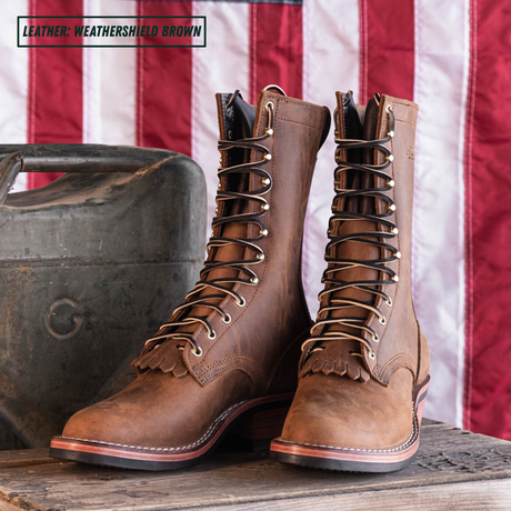 A pair of tall lace-up leather boots in a weathered brown finish, displayed on a wooden surface next to a vintage metal container with an American flag backdrop. A text overlay in the top-left corner reads 'LEATHER: WEATHERSHIELD BROWN'.