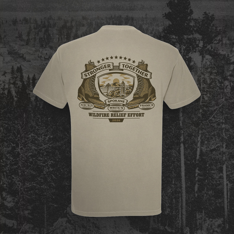 A light beige crewneck t-shirt shown from the back, featuring a large brown and gold graphic print with the text 'Stronger Together Wildfire Relief Effort' and 'Spokane County, Washington'. The shirt is displayed against a dark, moody forest background.