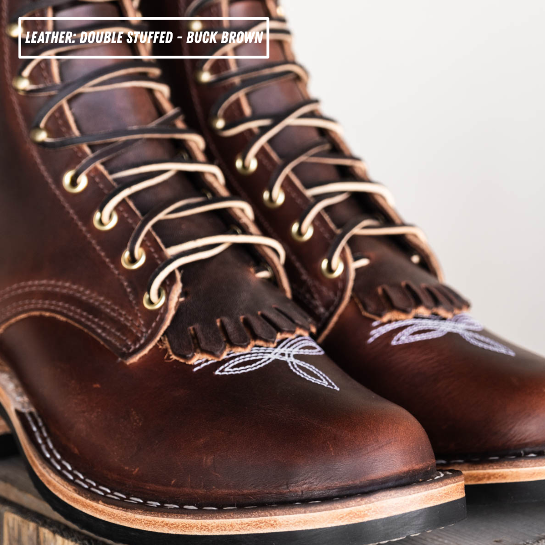 A close-up of a pair of double-stuffed buck brown leather lace-up boots with fringe detailing and white embroidered patterns near the toes, shown on a neutral background with a text overlay at the top.