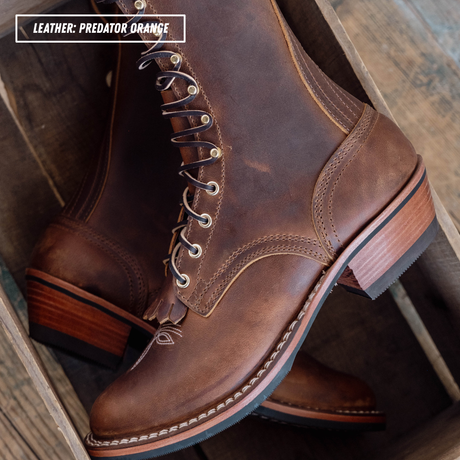 A pair of brown leather lace-up boots with stacked wooden heels, displayed inside a rustic wooden crate. A text overlay in the top left reads 'LEATHER: PREDATOR ORANGE'.