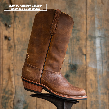 A single brown leather cowboy boot with a distressed finish, shown standing on a display stand against a rustic wooden plank background. A text overlay in the top-left corner reads 'LEATHER: PREDATOR ORANGE / SHRUNKEN BISON BROWN'.