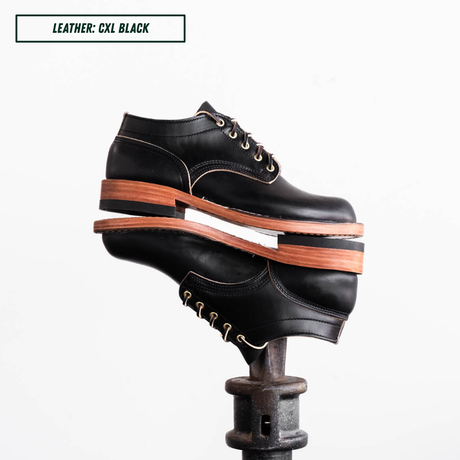 A pair of black leather lace-up shoes stacked on a metal stand against a white background, with a text label in the top-left corner reading 'LEATHER: CXL BLACK'.