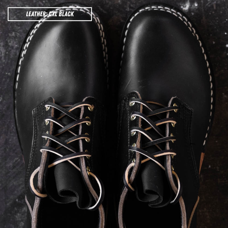 A pair of black leather lace-up boots shown from a top-down view on a dark textured surface. White contrast stitching outlines the edges, and the laces are threaded through brass eyelets.