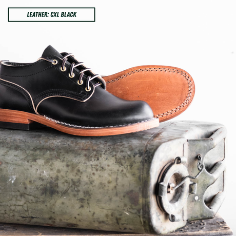 A black leather lace-up shoe shown from the side and top, resting on a vintage metal box. The image includes a text label in the top-left corner that reads 'LEATHER: CXL BLACK'.