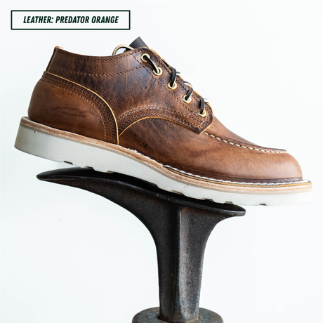 A single brown leather lace-up shoe displayed on a black metal stand against a white background, with a text label in the top-left corner reading 'LEATHER: PREDATOR ORANGE'.