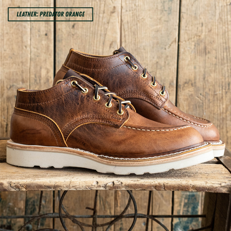 A pair of brown leather lace-up ankle boots with white soles, displayed on a rustic wooden bench against a wood plank wall. A text overlay in the top left reads 'LEATHER: PREDATOR ORANGE'.