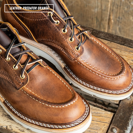 A pair of brown leather lace-up boots with white contrast stitching and white soles, photographed on a rustic wooden surface. A text overlay in the top left reads 'LEATHER - PREDATOR ORANGE'.