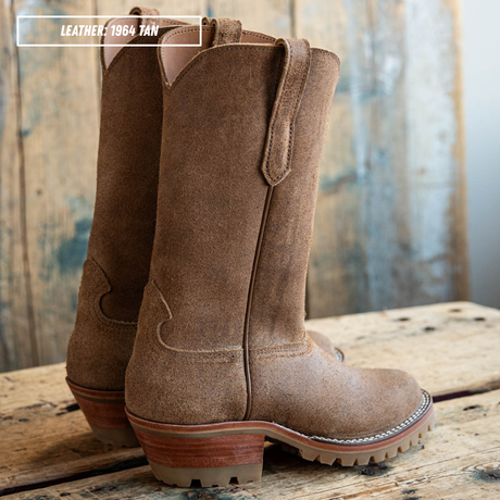 A pair of mid-calf western boots in tan leather, shown standing on a rustic wooden plank with a weathered wood background. A text overlay in the top left reads 'LEATHER - 1984 TAN'.