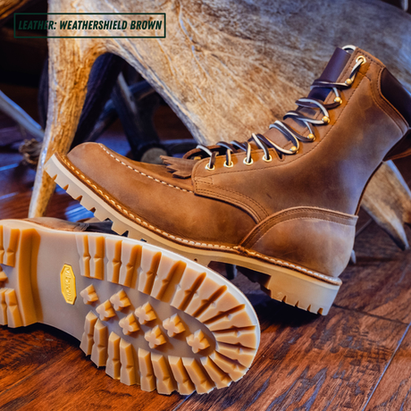 A pair of brown leather lace-up boots with fringed detailing, shown on a rustic wooden floor with a large antler prop. One boot is standing upright while the other lies on its side to display the textured off-white sole and a gold heel badge.