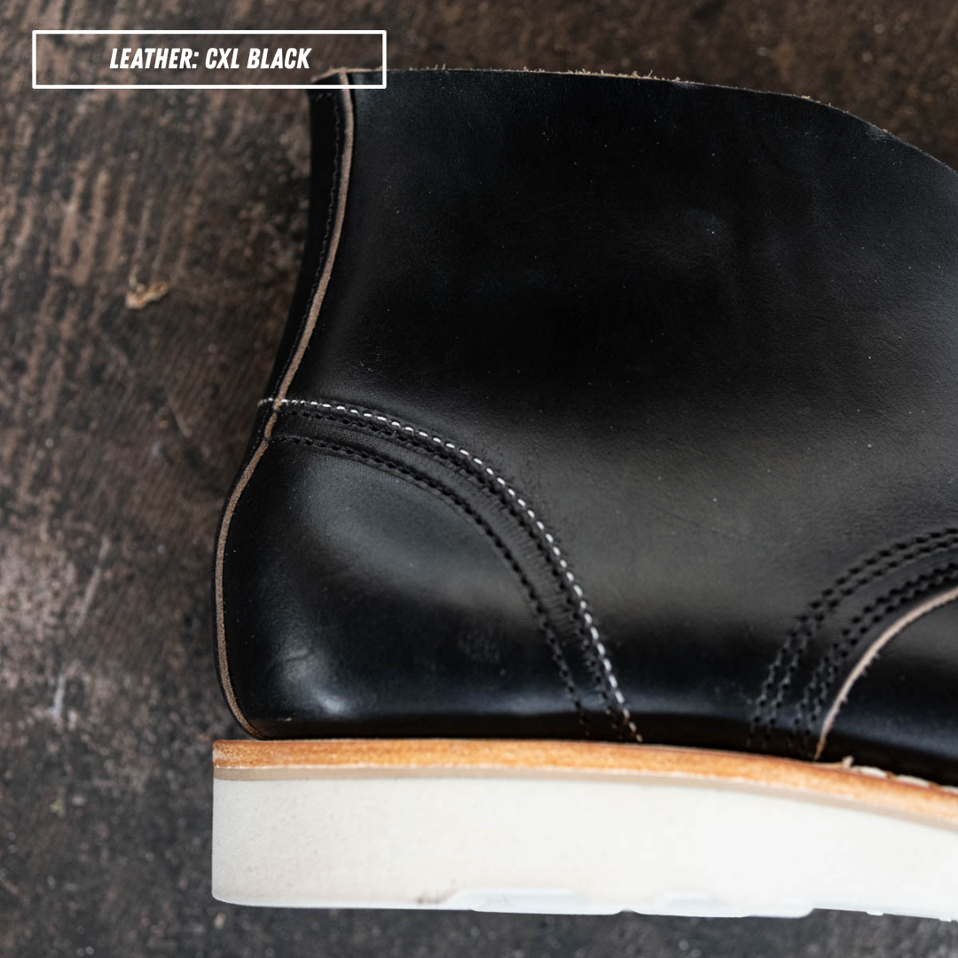 detail shot of a black leather ankle boot with a light beige sole, shown in a side profile on textured concrete. Zoomed in on internal heel counter. A text box in the top left corner reads 'LEATHER: CXL BLACK'.