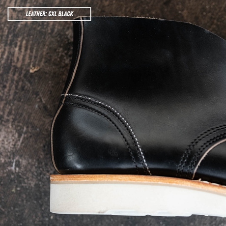 detail shot of a black leather ankle boot with a light beige sole, shown in a side profile on textured concrete. Zoomed in on internal heel counter. A text box in the top left corner reads 'LEATHER: CXL BLACK'.