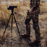 A person in camouflage outdoor clothing stands beside a black binocular mounted on a tripod in a dry grassy field. A text box in the top left reads 'LEATHER: WEATHERSHIELD BROWN'.