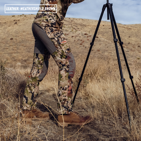 A person wearing camouflage hunting pants with grey knee panels and brown leather boots stands in a dry grassy field next to a tripod. A text overlay in the top left reads 'LEATHER: WEATHERSHIELD BROWN'.