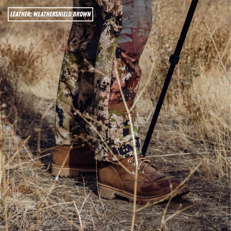 A close-up outdoor shot of a person wearing camouflage pants and brown leather boots, standing in dry grass with a black hiking pole visible. Text in the top-left reads 'LEATHER: WEATHERSHIELD BROWN'.