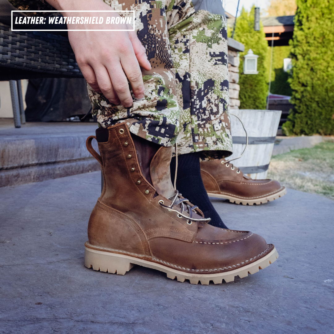 A person wearing brown leather lace-up boots with a rugged off-white sole, standing outdoors on a concrete surface. The pants are camouflage, and a hand is pulling the leg up to show the boot opening.