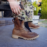 A person wearing brown leather lace-up boots with a rugged off-white sole, standing outdoors on a concrete surface. The pants are camouflage, and a hand is pulling the leg up to show the boot opening.