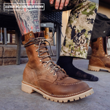 A close-up of a person wearing a brown leather lace-up work boot, with another boot visible in the background and patterned pants. A text overlay in the top left reads 'LEATHER WEATHERSHIELD BROWN'.