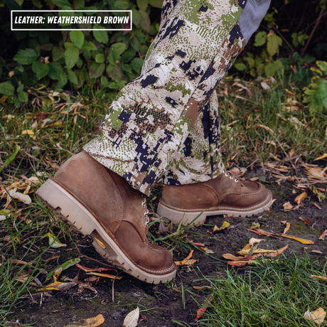 A person wearing camouflage pants and brown leather work boots standing outdoors on grass and dirt with scattered leaves. A text box in the top left reads 'LEATHER: WEATHERSHIELD BROWN'.