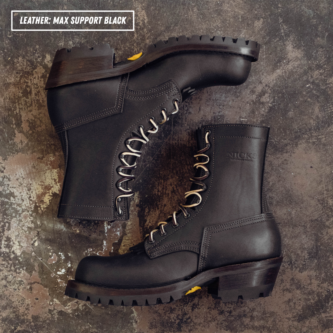 Two black leather work boots arranged on a textured dark surface. One boot is positioned upright and the other is angled on top, showing the side, toe, and laces.