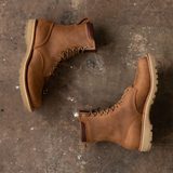A pair of tan leather lace-up boots with thick beige soles, arranged on a textured dark brown rustic surface.