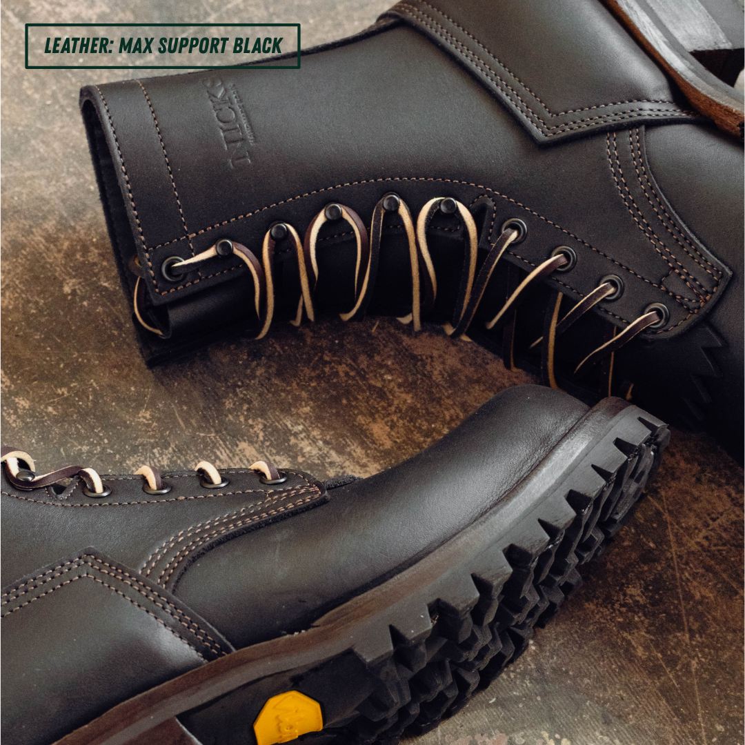 Close-up of a pair of black leather lace-up boots on a rustic brown textured surface. One boot is angled to show the side profile with metal eyelets and laces.