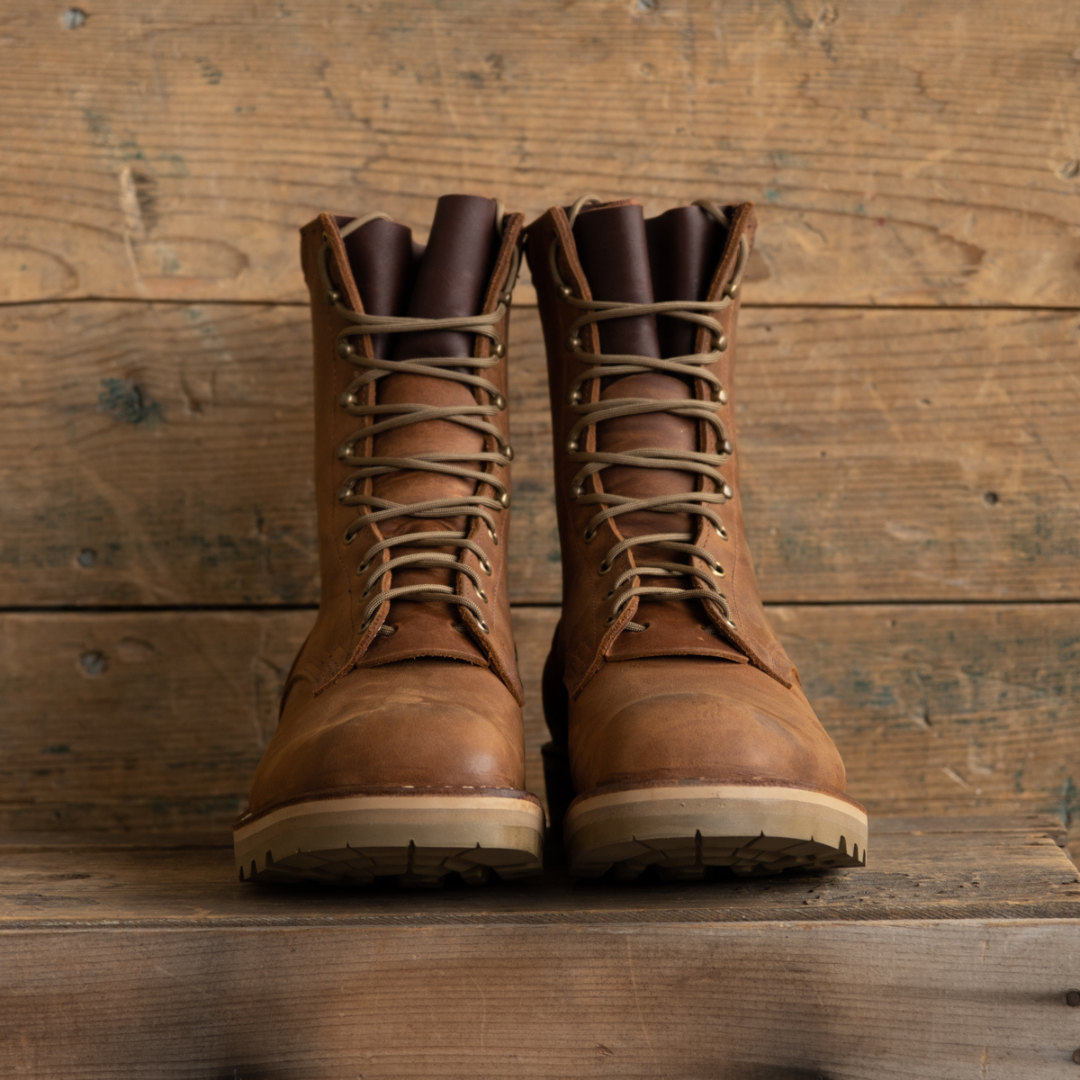 A pair of brown leather lace-up work boots with tan laces and light beige soles, displayed on a rustic wooden surface against a wooden plank background.