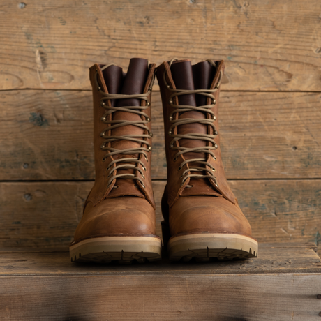 A pair of brown leather lace-up work boots with tan laces and light beige soles, displayed on a rustic wooden surface against a wooden plank background.