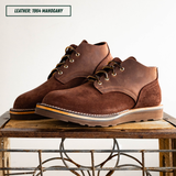 A pair of dark brown suede and leather chukka-style boots with white stitching and metal eyelets, displayed on a rustic wooden table against a plain white background. A text label in the top left reads 'LEATHER: 1964 MAGOGANY'.