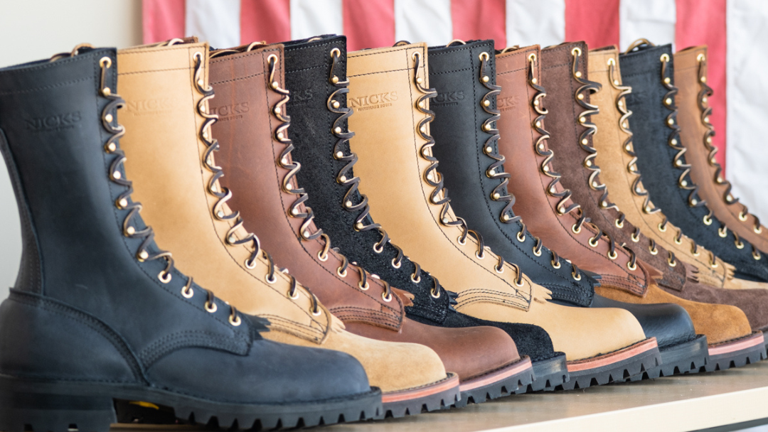 A row of lace-up work boots in multiple colors including black, tan, brown, and charcoal, lined up on a shelf with an American flag hanging in the background.