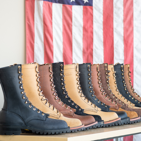 A row of lace-up work boots in multiple colors including black, tan, brown, and charcoal, lined up on a shelf with an American flag hanging in the background.
