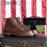 A pair of brown leather lace-up boots displayed on a vintage suitcase in front of a red and white striped flag backdrop, with a black case and a Remington Express box behind them. A label in the top left reads 'LEATHER: 1964 BROWN'.