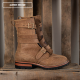 A pair of mid-calf leather boots in tan, featuring multiple strap details with brass-tone buckles and a rugged lug sole, displayed on a rustic wooden surface with a wooden background.