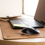 A black and white computer mouse resting on a light brown leather desk mat, with a laptop and notebook in the background near a bright window.