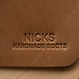 Close-up of a brown leather boot showing the embossed brand name 'NICKS HANDMADE BOOTS' on a light wood surface.