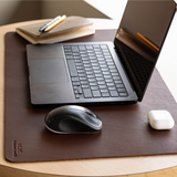 A dark brown leather desk mat on a light wood table, styled with a black laptop, a black mouse, a white earbud case, and a notebook with a pen.
