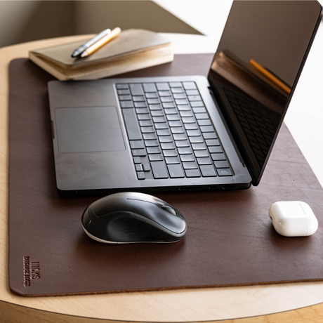 A dark brown leather desk mat on a light wood table, styled with a black laptop, a black mouse, a white earbud case, and a notebook with a pen.