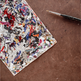 A square palette card covered in multicolored paint splatters, placed diagonally on a brown textured surface next to a paintbrush.