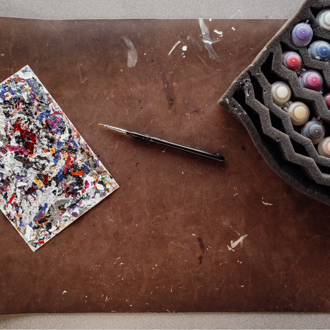 A flat-lay view of an artist's palette setup on a brown leather mat, featuring a paint-splattered palette card, a fine detail brush, and a foam tray holding multiple paint tubes.
