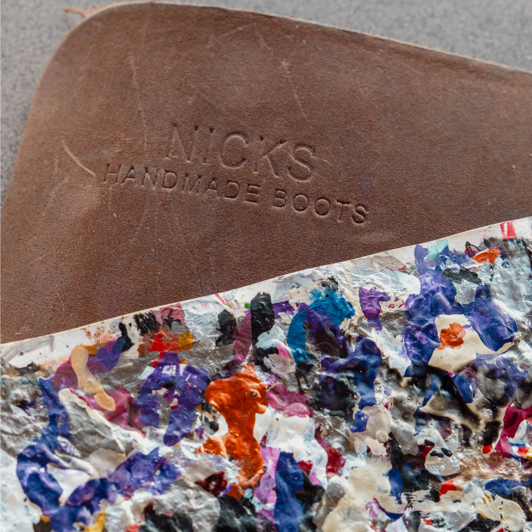 Close-up of a brown leather boot sole embossed with 'NICKS HANDMADE BOOTS' over a highly textured, multicolored abstract paint-splattered surface.